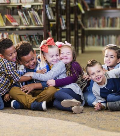 Six children sit on the floor of a library, laughing and hugging