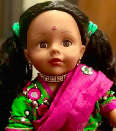 Doll Provides Representation 18 inch doll stands in a green a pink dress with pig tails in her hair and a bindi on her forehead.