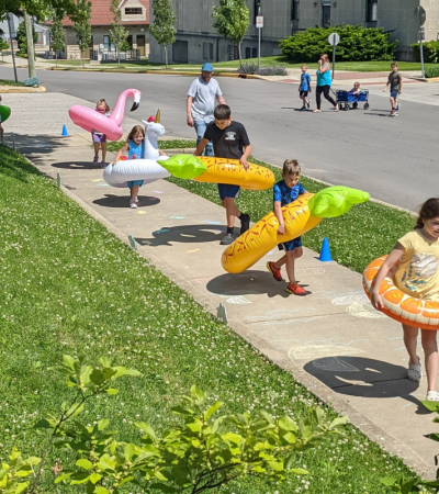 Photograph shows children outside playing a sidewalk chalk game with pool floaties on.