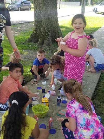 A girl in a pink dress standing among a group of kids sitting on the sidewalk making slime