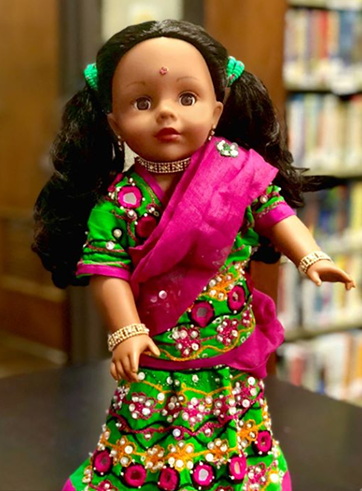 18 inch doll stands in a green a pink dress with pig tails in her hair and a bindi on her forehead.