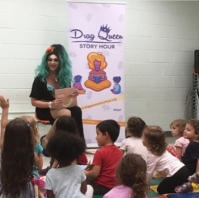 Drag Queen sits in front of audience of children holding a book smiling. A child raises their hand to ask a question. A poster reads "Drag Queen Story Hour" in the background.