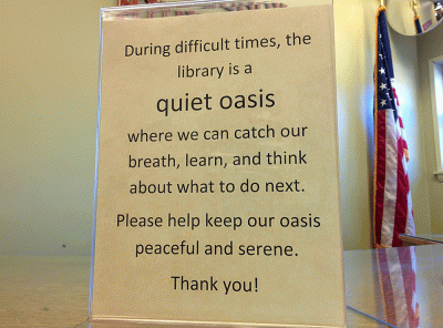 Yellow paper sign in plastic holder. Sign reads: "During difficult times, the library is a quiet oasis where we can catch our breath, learn, and think about what to do next. Please help keep our oasis peaceful and serene. Thank you!