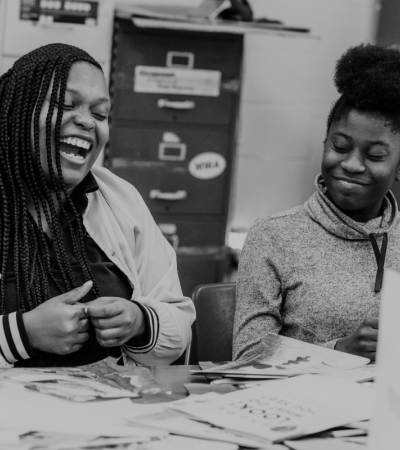 Black and white photograph shows two teens laughing at a table with collage materials.
