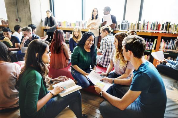 Photograph shows a group of college students sitting and talking in a library.