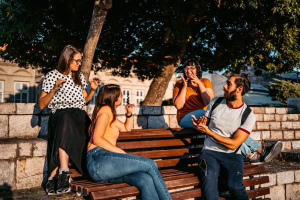 Photograph of a group of four friends sitting outside on a bench. They are facing each other and signing.