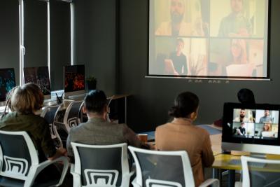 Three people seated in front of projector screen with a Zoom meeting screen pulled up with four people included.