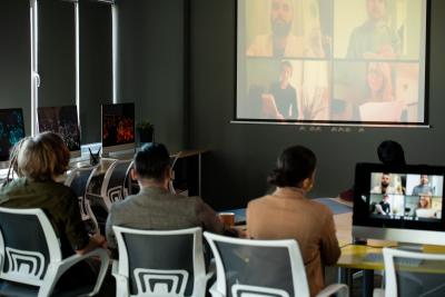 Three people seated in front of projector screen with a Zoom meeting screen pulled up with four people included. 